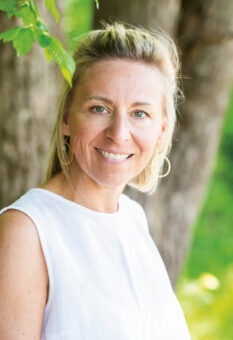 Eine Frau mit blondem Haar und einem weißen ärmellosen Oberteil lächelt, während sie im Freien vor einem Baum steht, mit grünem Laub im Hintergrund.