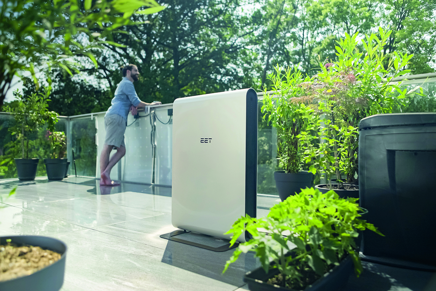 Ein Mann steht auf einem Balkon mit Glasgeländer, umgeben von Topfpflanzen und Grünzeug. Im Vordergrund ist ein weißes Gerät mit der Aufschrift "EET" zu sehen. Im Hintergrund sind Bäume an einem sonnigen Tag zu sehen.
