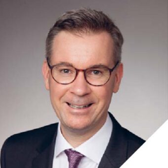 Ein Mann mit Brille, dunklem Anzug, weißem Hemd und gemusterter Krawatte lächelt in die Kamera vor einem schlichten, hellen Hintergrund.