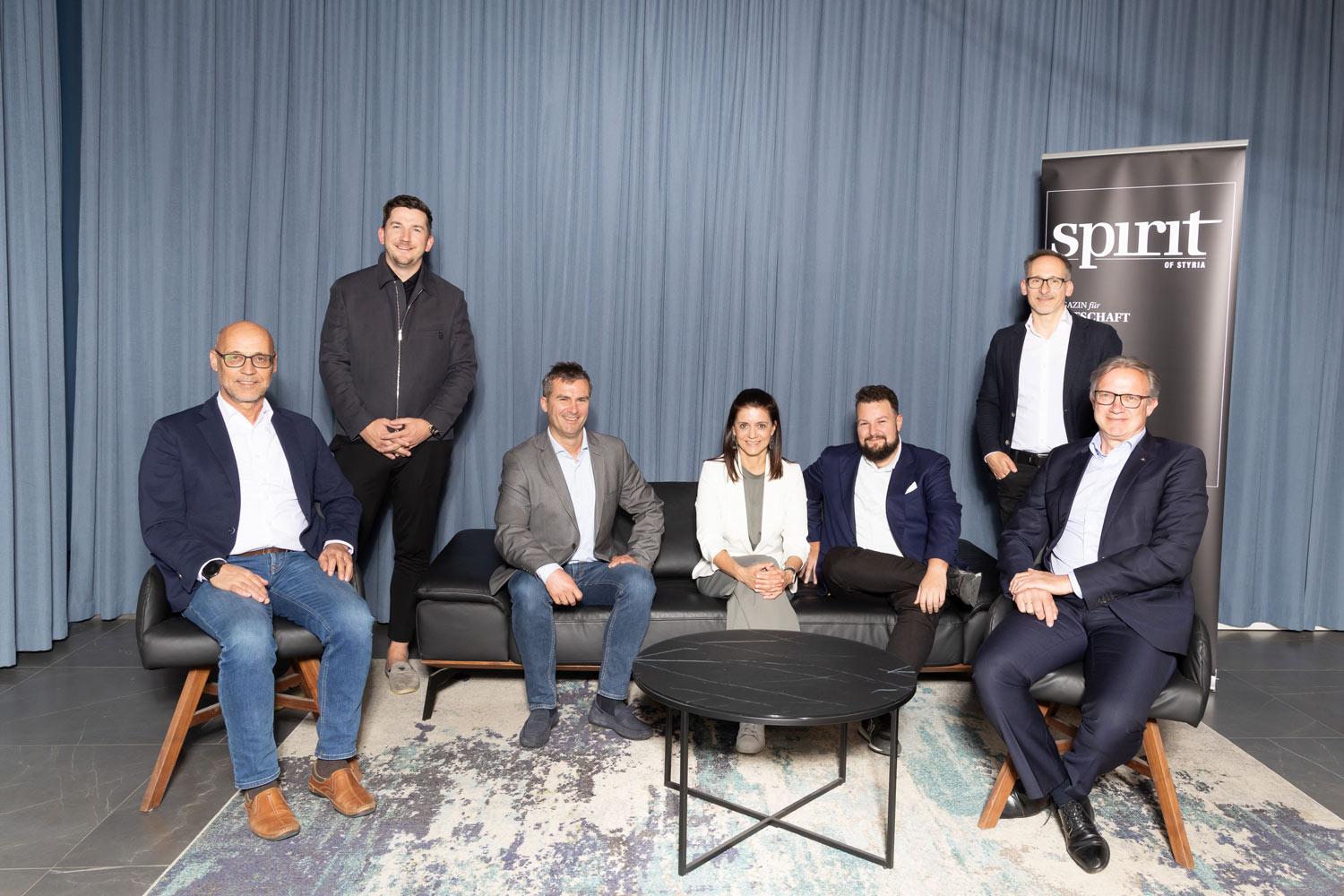 Sieben professionell gekleidete Personen posieren vor einem blauen Vorhang, wobei vier auf einem Sofa sitzen und drei dahinter stehen. In der Szene sind ein schwarzer runder Tisch und ein "Spirit"-Schild zu sehen.