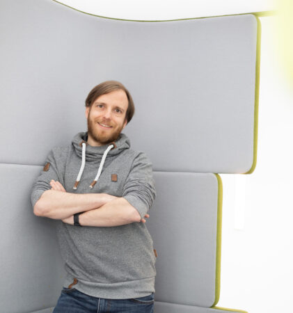 Ein Mann mit Bart, grauem Kapuzenpulli und Jeans, steht mit verschränkten Armen da und lächelt in die Kamera. Er lehnt an einer hellgrauen, gepolsterten Wand in einem hellen, modernen Raum.