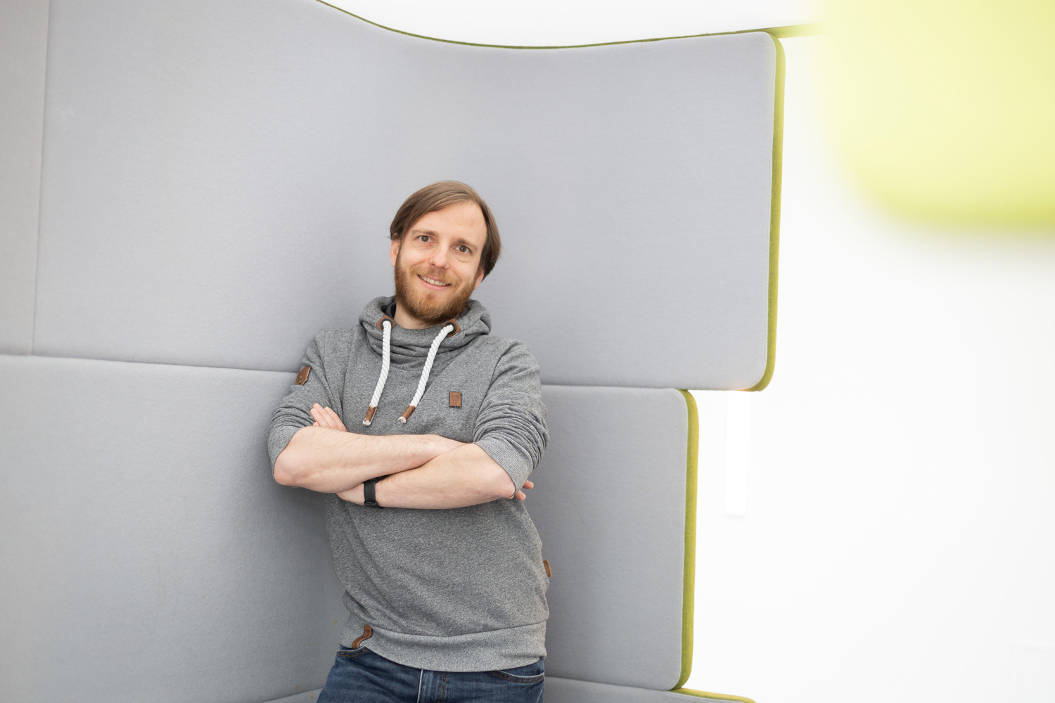 Ein Mann mit Bart, grauem Kapuzenpulli und Jeans, steht mit verschränkten Armen da und lächelt in die Kamera. Er lehnt an einer hellgrauen, gepolsterten Wand in einem hellen, modernen Raum.