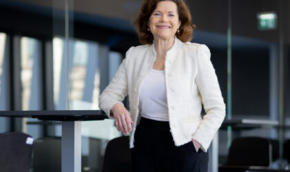 Eine Frau mit schulterlangen braunen Haaren, die einen weißen Blazer und eine schwarze Hose trägt, steht lächelnd neben einem hohen Tisch in einem modernen Büro mit Glaswänden und leeren Stühlen im Hintergrund.