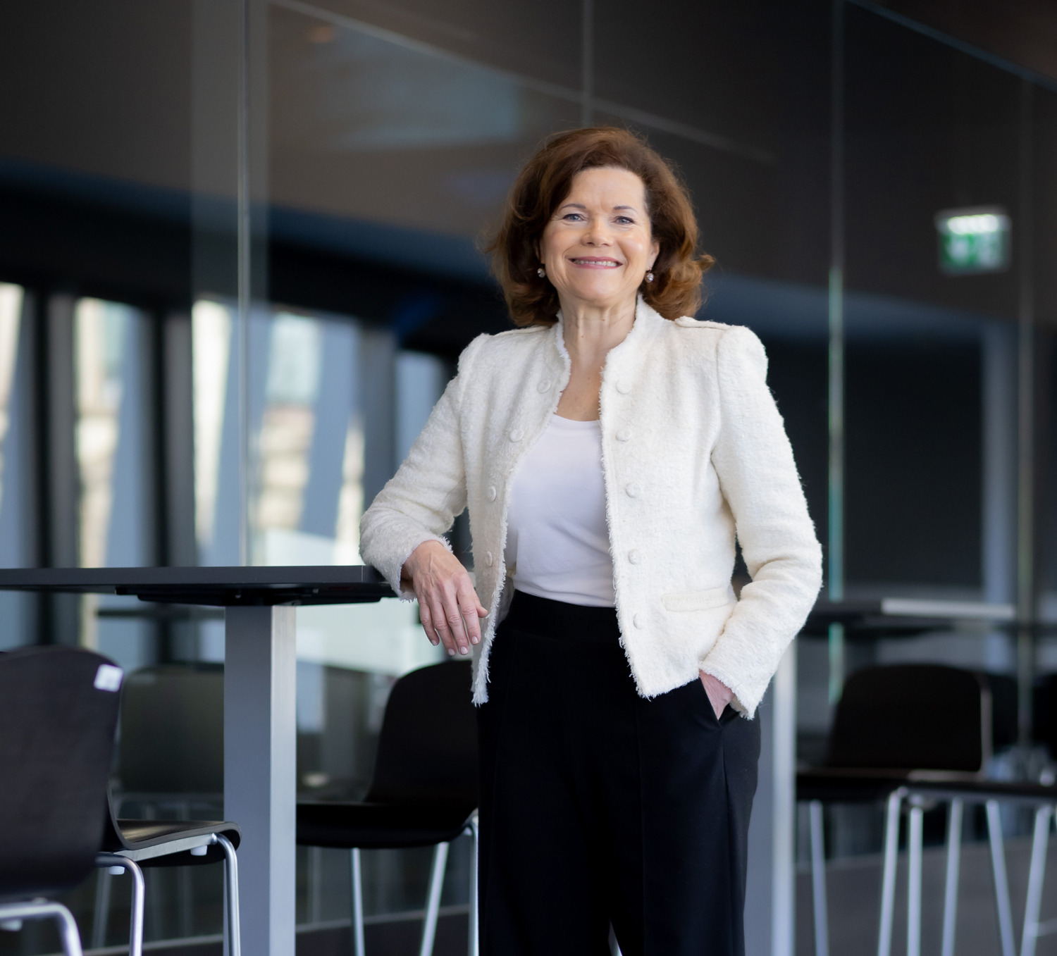 Eine Frau mit schulterlangen braunen Haaren, die einen weißen Blazer und eine schwarze Hose trägt, steht lächelnd neben einem hohen Tisch in einem modernen Büro mit Glaswänden und leeren Stühlen im Hintergrund.