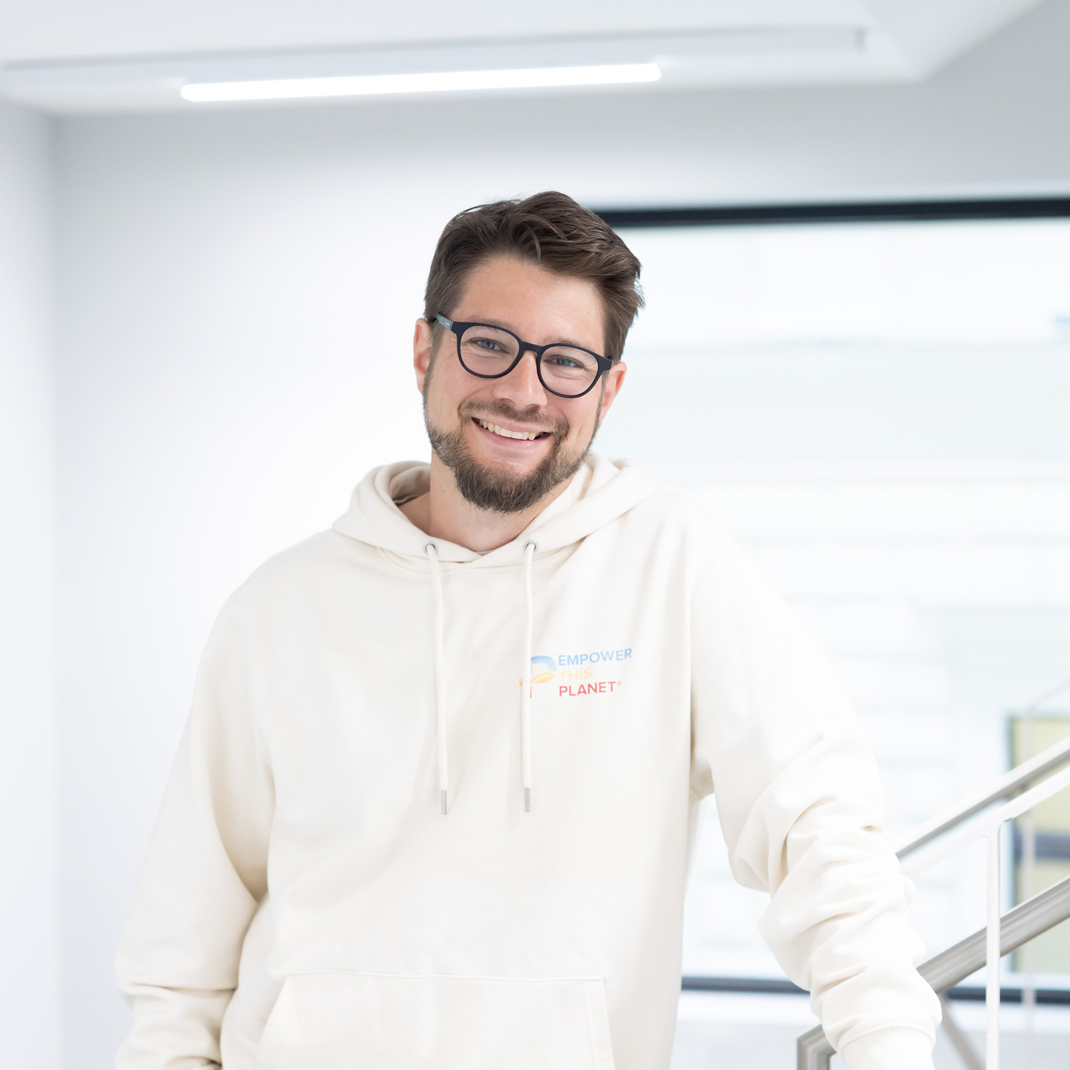 Ein Mann mit Brille und Bart, der einen hellen Kapuzenpulli mit der Aufschrift "Empower Planet" trägt, lächelt, während er in einem Haus neben einem Metallgeländer steht. Der Hintergrund ist hell und modern.