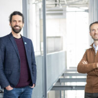 Zwei Männer stehen in einem Innenraum eines hellen, modernen Gebäudes. Der eine hat lockiges Haar und einen Bart und trägt einen marineblauen Blazer, der andere hat kurze Haare, einen Bart und einen braunen Pullover mit Reißverschluss und steht mit verschränkten Armen da.