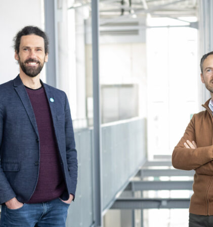 Zwei Männer stehen in einem Innenraum eines hellen, modernen Gebäudes. Der eine hat lockiges Haar und einen Bart und trägt einen marineblauen Blazer, der andere hat kurze Haare, einen Bart und einen braunen Pullover mit Reißverschluss und steht mit verschränkten Armen da.
