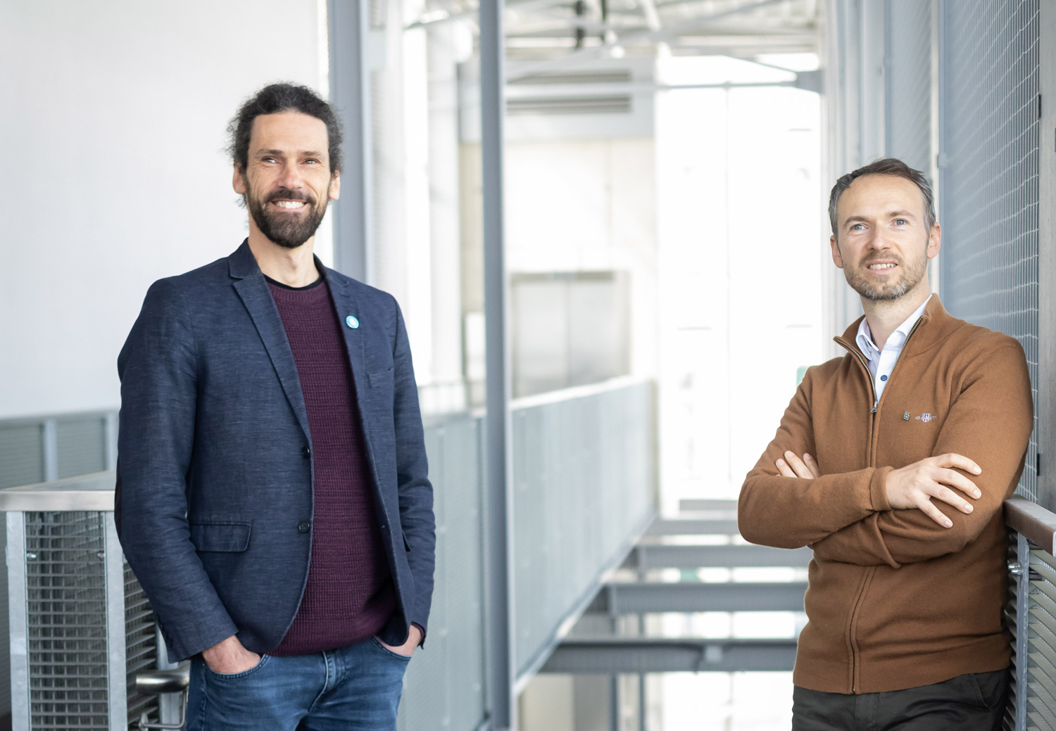 Zwei Männer stehen in einem Innenraum eines hellen, modernen Gebäudes. Der eine hat lockiges Haar und einen Bart und trägt einen marineblauen Blazer, der andere hat kurze Haare, einen Bart und einen braunen Pullover mit Reißverschluss und steht mit verschränkten Armen da.