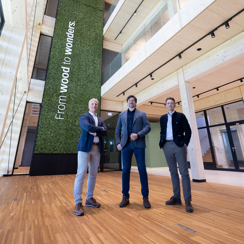 Drei Männer in Geschäftskleidung stehen selbstbewusst in einem geräumigen, modernen Holzgebäude mit hohen Decken; eine grüne Wand hinter ihnen zeigt den Text "From wood to wonders".