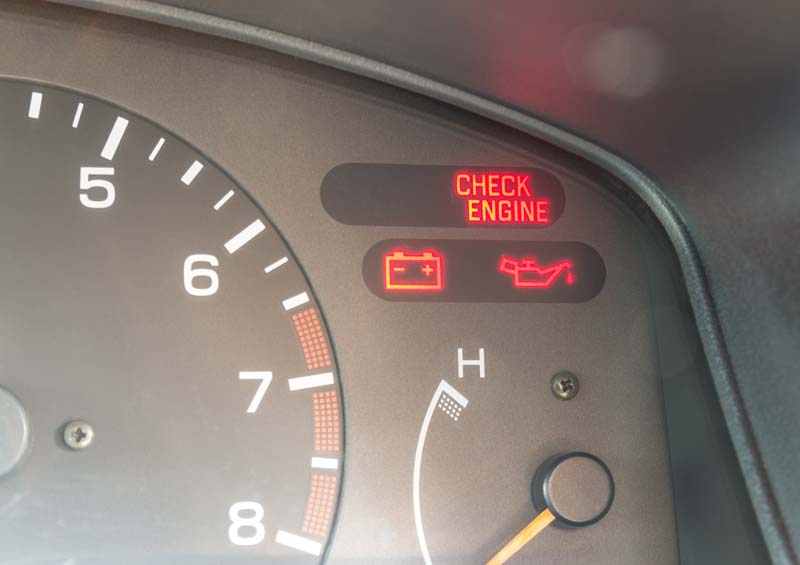 Nahaufnahme eines Autoarmaturenbretts mit beleuchteten Warnleuchten: "Motor prüfen", Batterie- und Öldruckanzeige neben dem Tachometer und der Temperaturanzeige.