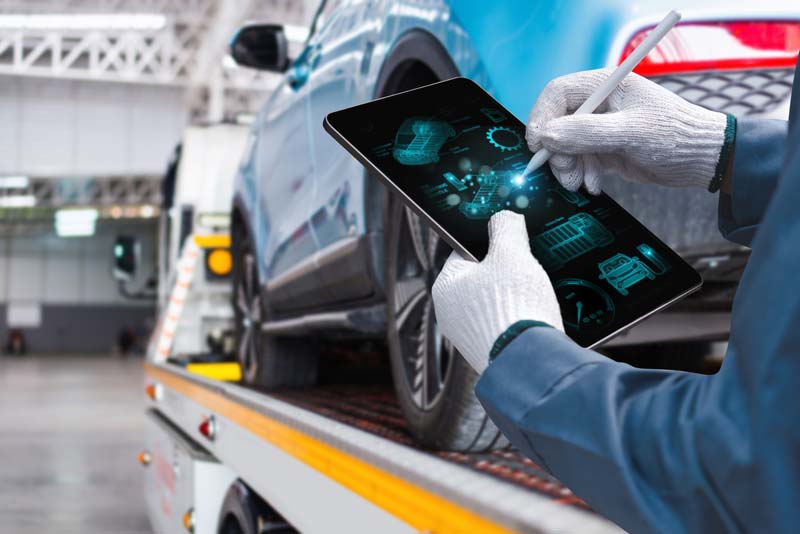 Eine Person mit Handschuhen benutzt einen Stift auf einem Tablet, das digitale Autodiagnosen anzeigt, und steht neben einem blauen Auto auf einer Plattform in einer hellen Industriehalle.