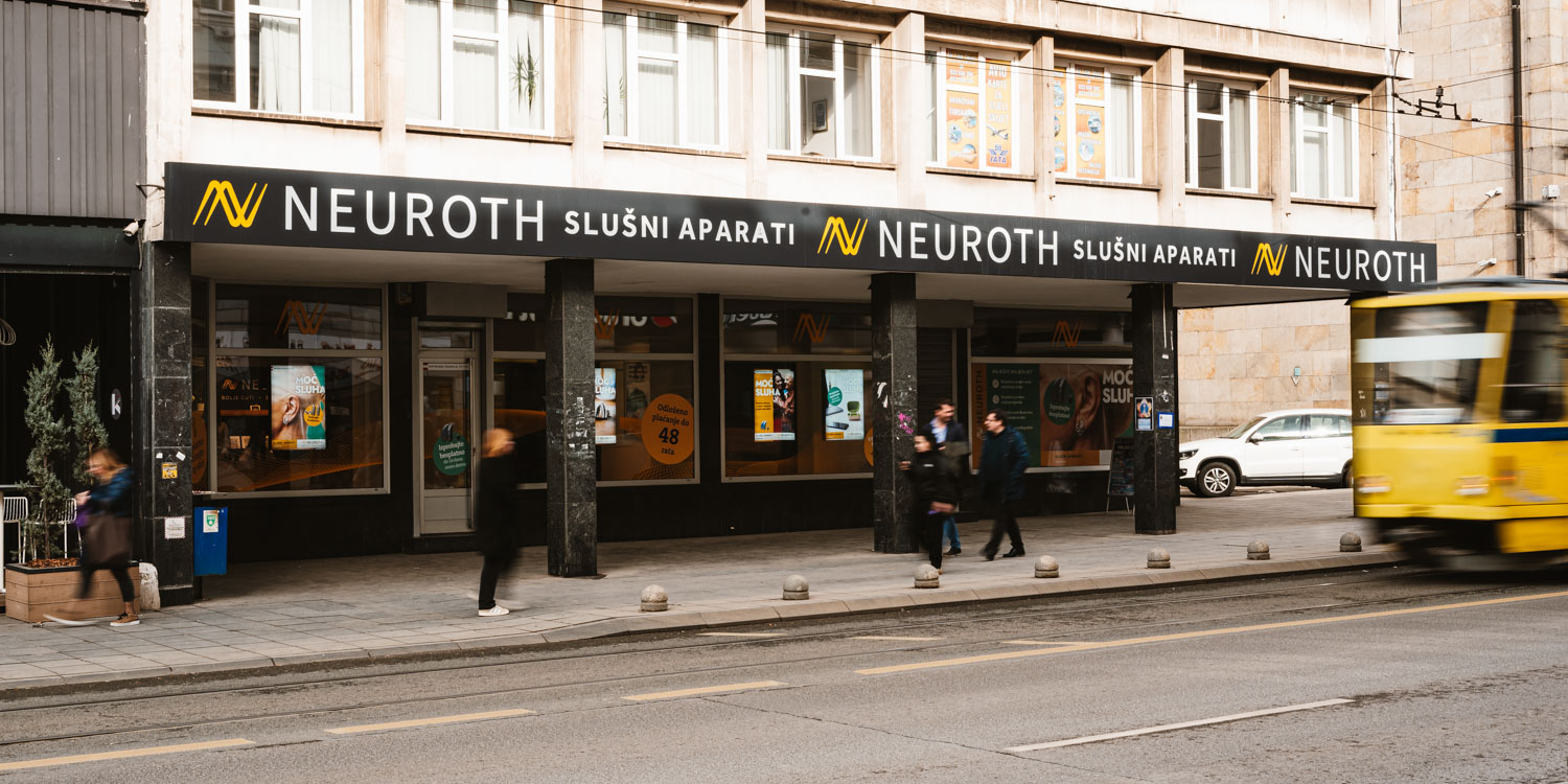 Straßenansicht eines NEUROTH-Hörgeräteladens mit mehreren vorbeilaufenden Personen und einer gelben Straßenbahn auf der rechten Seite des Bildes. Große Schilder an der Fassade zeigen den Namen des Geschäfts in weißen Buchstaben.