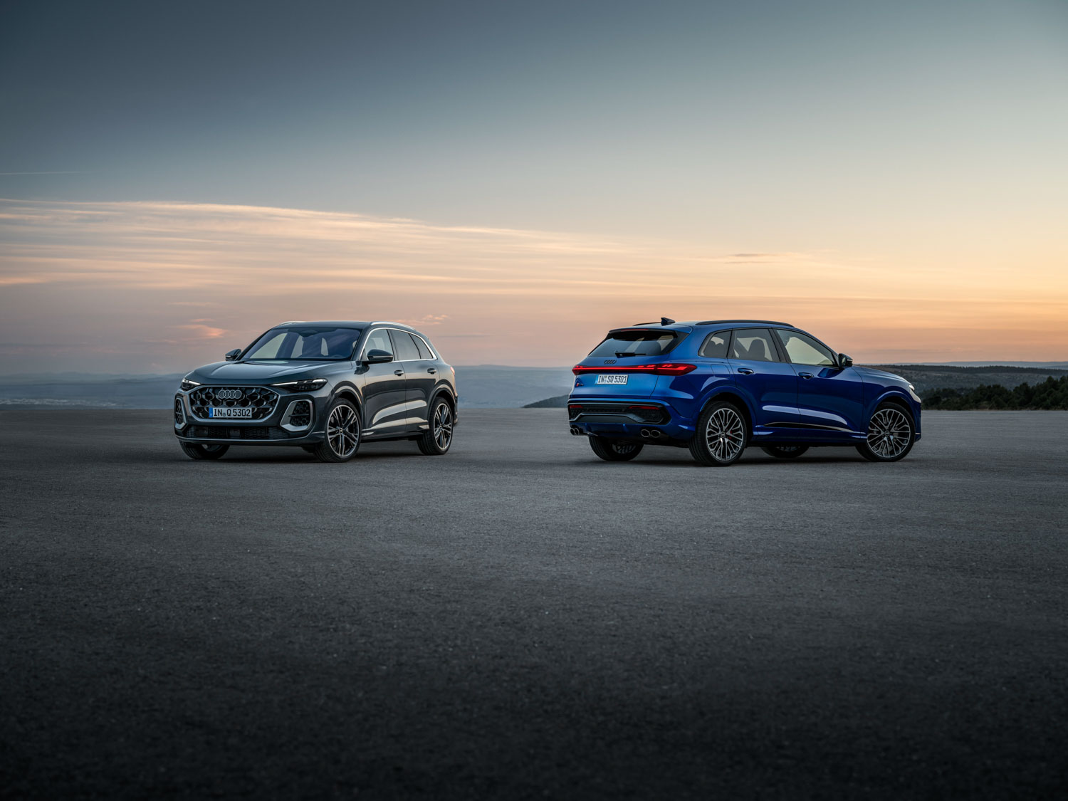 Zwei moderne Geländewagen, die bei Sonnenuntergang auf einer offenen, gepflasterten Fläche geparkt sind; ein silberner Audi mit Blick nach vorne und ein blauer Audi mit Blick nach hinten, mit Bergen und einem bunten Himmel im Hintergrund.