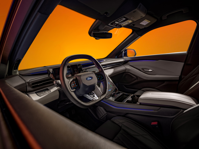 Innenansicht eines modernen Autos mit einem Ford-Lenkrad, einem digitalen Armaturenbrett und einer Mittelkonsole, mit schwarz-weißen Sitzen und einem durch die Fenster sichtbaren orangefarbenen Hintergrund.