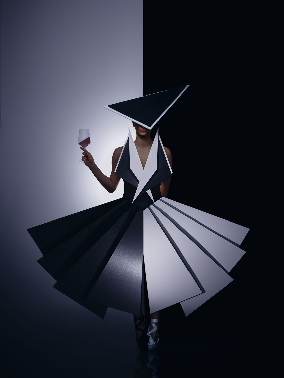 Eine Person in einem geometrischen schwarz-weißen Kleid und einem dreieckigen Hut steht vor einem geteilten schwarz-weißen Hintergrund und hält ein Glas Rotwein in der Hand. Das gewagte Outfit zeichnet sich durch scharfe, eckige Formen und dramatische Kontraste aus.