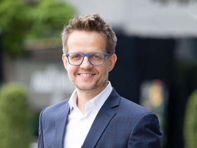 Ein Mann, der eine Brille und eine dunkle Anzugsjacke über einem weißen Hemd trägt, lächelt in die Kamera. Der Hintergrund ist im Freien mit Grün und unscharfen Gebäuden.