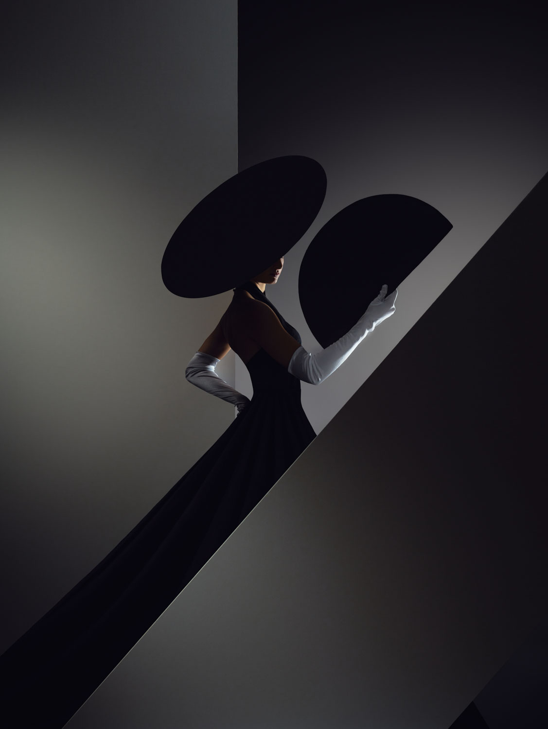 Eine Person in einem dramatischen schwarzen Kleid, einem breitkrempigen Hut und weißen Handschuhen posiert hinter geometrischen Formen und hält einen großen schwarzen Fächer in einer schwach beleuchteten, minimalistischen Umgebung.
