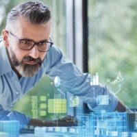 Ein Mann mit Brille und Bart schaut aufmerksam auf eine digitale Schnittstelle, auf der blaue holografische Diagramme und Schaubilder angezeigt werden, die eine Analyse oder Planung suggerieren, während durch ein Fenster im Hintergrund grüne Bäume zu sehen sind.