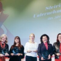 Sieben Frauen stehen auf der Bühne, halten Preise in der Hand und lächeln für ein Gruppenfoto. Hinter ihnen erscheint auf einer Leinwand der Schriftzug "Steirische Unternehmerin des Jahres 2024" zusammen mit einer stilisierten Grafik einer Frau.