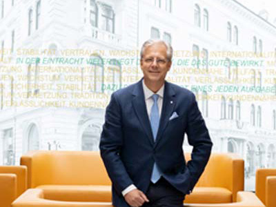 Ein Mann im Anzug steht vor einer orangefarbenen Couch und lächelt. Hinter ihm befindet sich eine helle Wand mit Worten in verschiedenen Farben und ein großes Fenster, das ein historisches Gebäude zeigt.