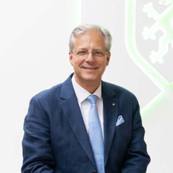 Ein Mann mittleren Alters mit grauem Haar und Brille, der einen blauen Anzug, ein weißes Hemd und eine hellblaue Krawatte trägt, lächelt und posiert vor einem hellen Hintergrund mit einem teilweise sichtbaren geometrischen Muster.