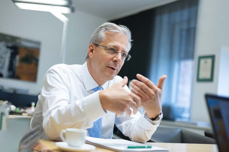 Ein Mann mittleren Alters mit weißem Hemd und blauer Krawatte gestikuliert, während er an einem Schreibtisch in einem Büro spricht, vor dem eine Kaffeetasse, Papiere und ein Stift liegen.