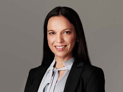Eine Frau mit langen dunklen Haaren, die einen schwarzen Blazer und eine helle Bluse mit einer Schleife im Nacken trägt, lächelt in die Kamera vor einem schlichten grauen Hintergrund.