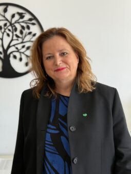 Eine Frau mit schulterlangen braunen Haaren, die einen schwarzen Blazer über einem blau gemusterten Oberteil trägt, steht in einem Innenraum vor einer weißen Wand mit einem dekorativen schwarzen Baummuster. Sie lächelt sanft in die Kamera.