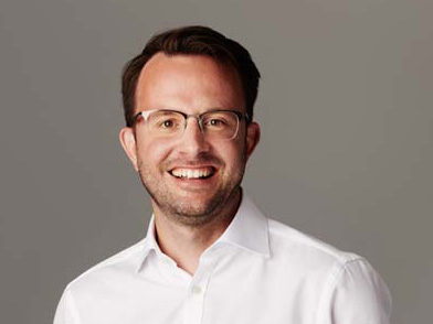 Ein Mann mit kurzen braunen Haaren und Brille, der ein weißes Hemd trägt, lächelt in die Kamera vor einem schlichten grauen Hintergrund.