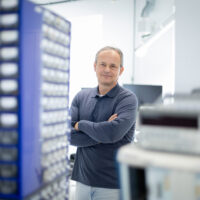 Ein Mann mit kurzen grauen Haaren steht mit verschränkten Armen in einem hellen, modernen Labor oder Arbeitsraum, umgeben von Regalen mit Kleinteilen und elektronischen Geräten, und schaut selbstbewusst in die Kamera.