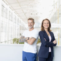 Zwei Personen stehen in einem Innenraum nebeneinander und lächeln selbstbewusst mit verschränkten Armen. Der Mann trägt ein weißes T-Shirt und hat Tattoos auf den Armen, die Frau trägt einen dunklen Anzug. Der Schauplatz ist ein helles, modernes Gebäude mit großen Fenstern.