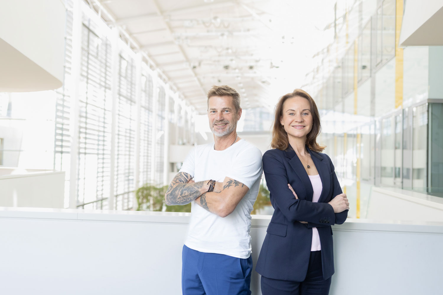 Zwei Personen stehen in einem Innenraum nebeneinander und lächeln selbstbewusst mit verschränkten Armen. Der Mann trägt ein weißes T-Shirt und hat Tattoos auf den Armen, die Frau trägt einen dunklen Anzug. Der Schauplatz ist ein helles, modernes Gebäude mit großen Fenstern.