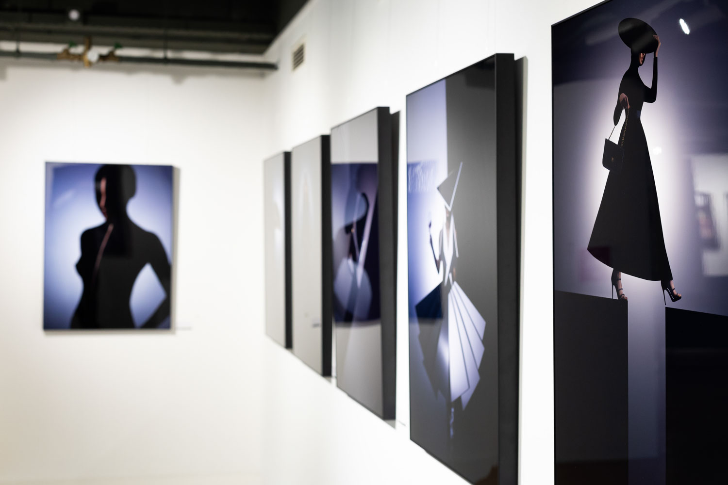 Eine Galeriewand zeigt gerahmte, stilisierte Silhouettenporträts von eleganten Frauen in modischen Posen mit minimalistischer Beleuchtung und Schatteneffekten. Der Fokus liegt auf vier Kunstwerken, ein weiteres ist im Hintergrund verschwommen.