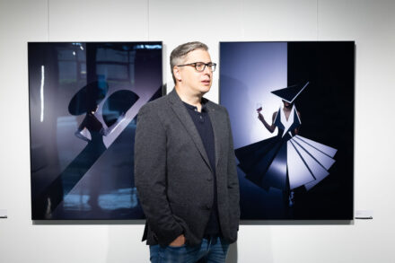 Ein Mann mit Brille und grauem Blazer steht in einer hell erleuchteten Galerie vor zwei modernen Kunstwerken, von denen das eine eine Figur mit einem Regenschirm und das andere eine geometrische Figur mit einem Glas zeigt.