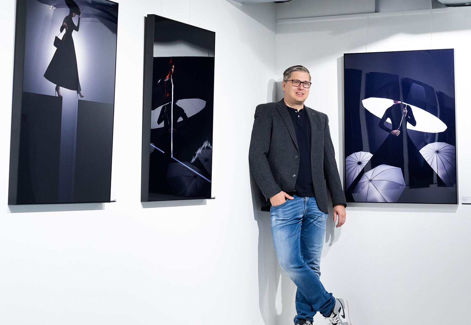 Ein Mann mit Brille, Blazer, Jeans und Turnschuhen lehnt an einer weißen Galeriewand neben drei ausgestellten modernen Modefotografien mit dunklem Thema.
