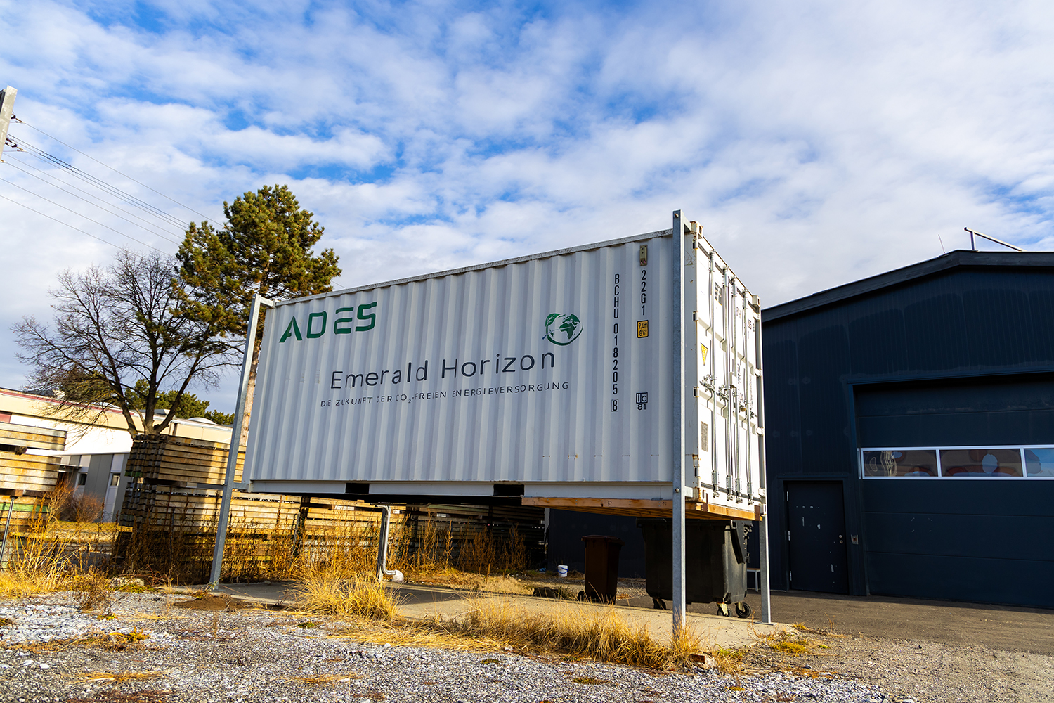 Ein weißer Schiffscontainer mit der Aufschrift ADES Emerald Horizon" steht auf Metallstützen neben einem dunkelblauen Gebäude. In der Nähe befinden sich Bäume und Palettenstapel unter einem teilweise bewölkten Himmel.