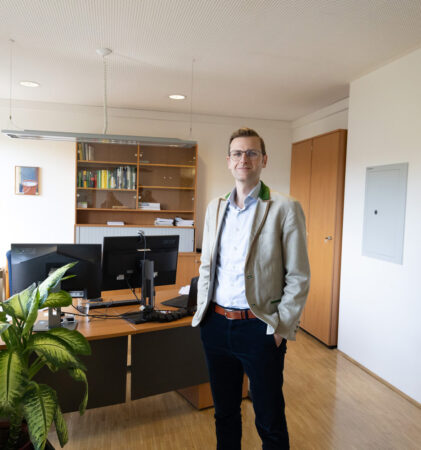 Ein Mann in einem beigen Blazer steht lächelnd in einem modernen Büro mit Holzboden, großen Fenstern, einem Schreibtisch mit Computermonitoren, Bücherregalen, Grünpflanzen und einer Landkarte an der Wand.