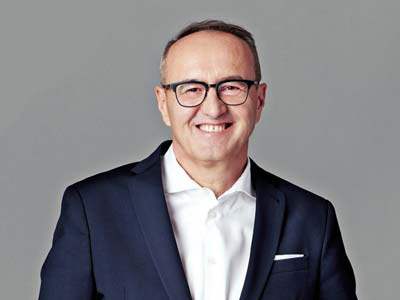 Ein Mann mit Brille, dunkler Anzugjacke und weißem Hemd lächelt in die Kamera vor einem schlichten grauen Hintergrund.
