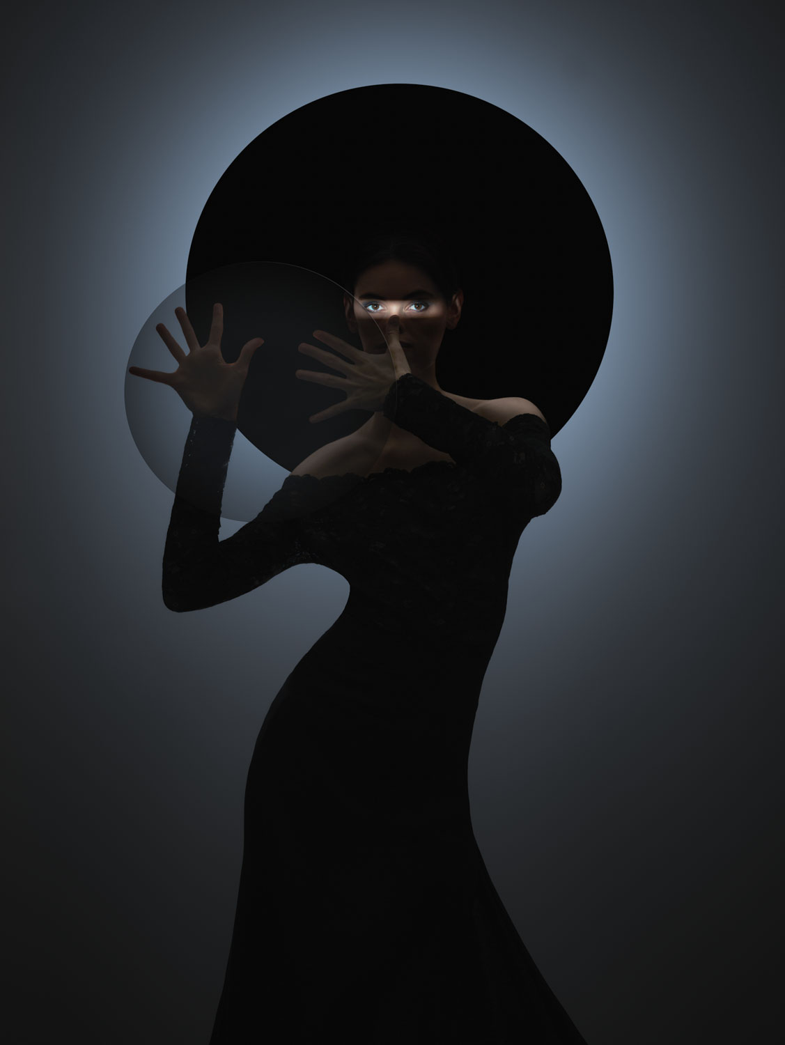 Eine Person in schwarzer, figurbetonter Kleidung und mit breitem Hut steht vor einem dunklen Hintergrund, mit leuchtenden Augen, die durch die Schatten hindurch sichtbar sind, und Händen, die vor einer durchscheinenden runden Form positioniert sind.