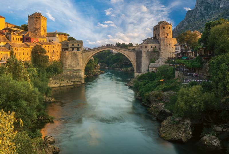 Eine malerische Steinbogenbrücke überspannt einen Fluss, umgeben von historischen Gebäuden und üppigem Grün unter einem blauen Himmel mit Wolken, wahrscheinlich bei Sonnenuntergang.