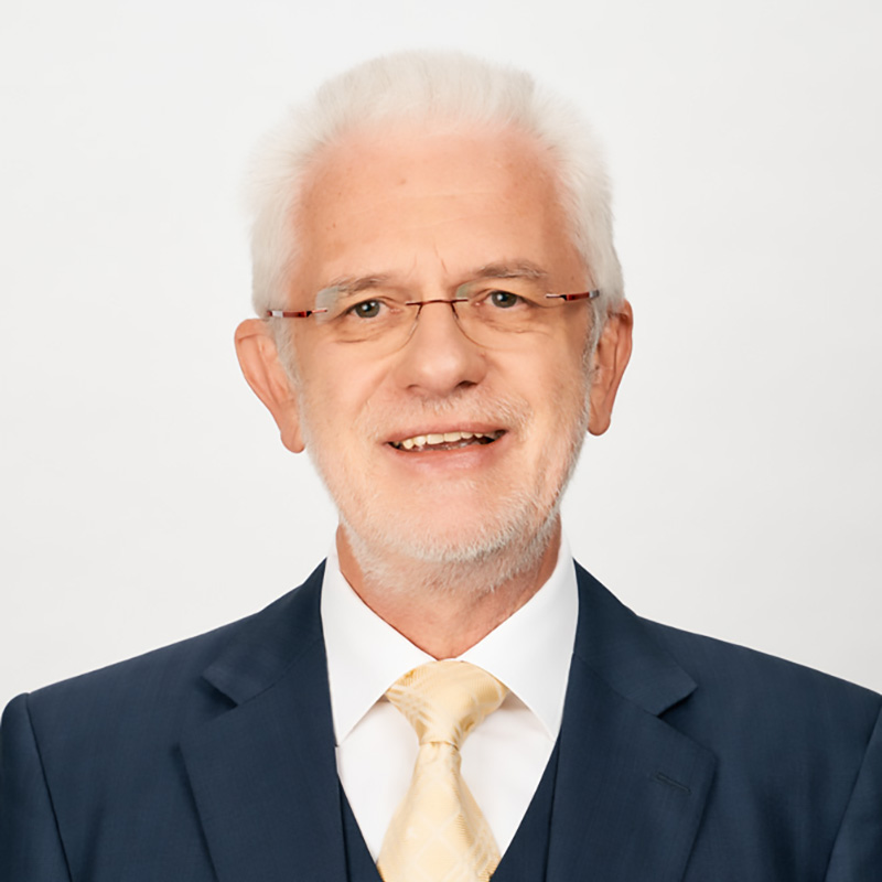 Ein älterer Mann mit kurzen weißen Haaren und Brille, der einen marineblauen Anzug, ein weißes Hemd und eine blassgelbe Krawatte trägt, lächelt vor einem schlichten weißen Hintergrund.