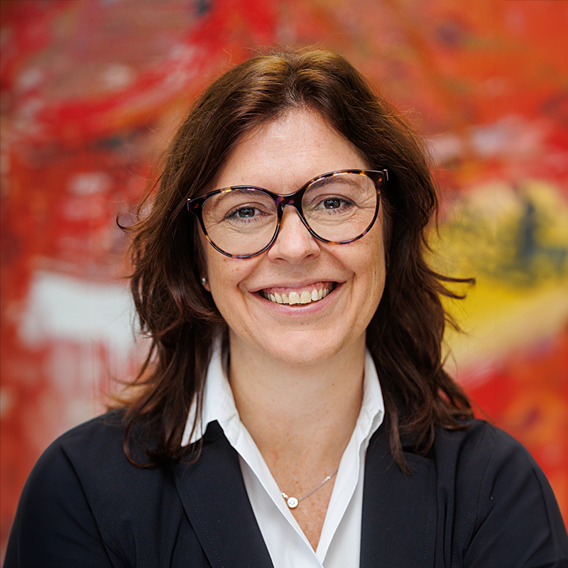 Eine Frau mit mittellangem braunem Haar, die eine Brille, eine weiße Bluse und einen dunklen Blazer trägt, lächelt vor einem bunten, verschwommenen roten und gelben Hintergrund.