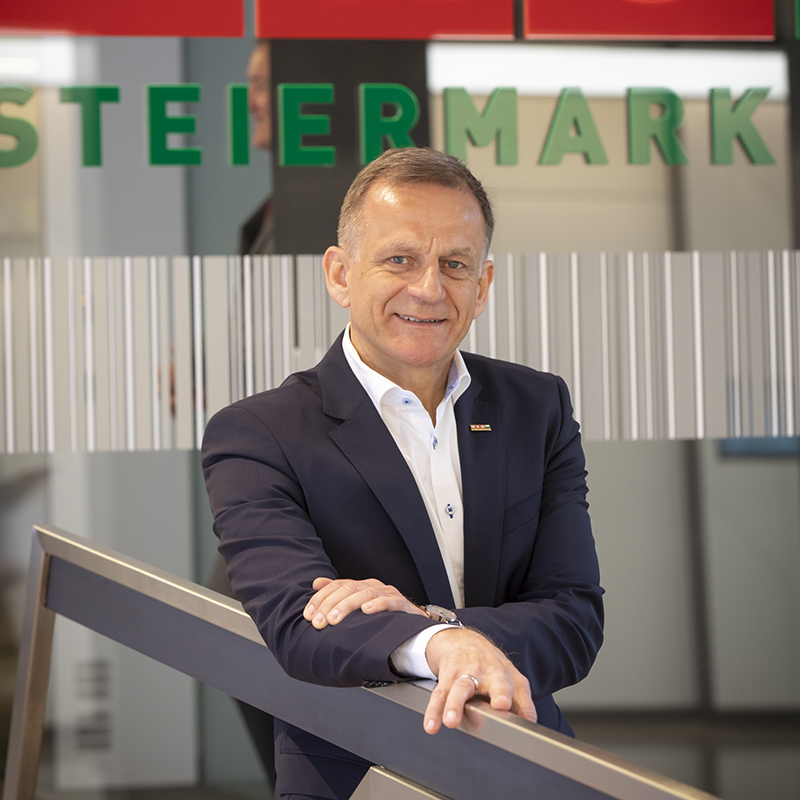 Ein Mann in einem dunklen Anzug und weißem Hemd lehnt an einem Geländer und lächelt in die Kamera. Hinter ihm ist in grüner Schrift auf Glas "STEIERMARK" zu lesen, im Hintergrund ist ein modernes Büro zu sehen.