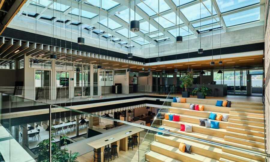 Modernes Büro mit Glasdecke, offenen Sitzgelegenheiten, Holzstufen mit bunten Kissen, großen Fenstern, Hängelampen, Gemeinschaftstischen und Grünpflanzen, die eine helle und geräumige Umgebung für die Zusammenarbeit schaffen.