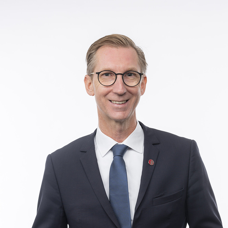Ein Mann mit Brille, dunklem Anzug, weißem Hemd und blauer Krawatte steht vor einem schlichten weißen Hintergrund und lächelt in die Kamera.