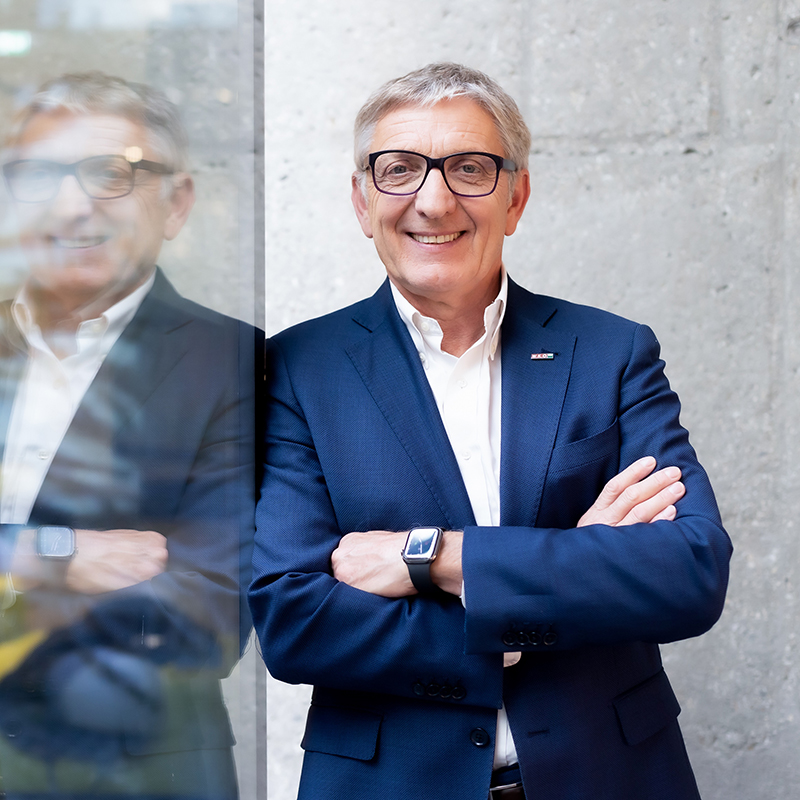 Ein älterer Mann in einem marineblauen Anzug und mit Brille steht mit verschränkten Armen lächelnd neben einer Glaswand, die sein Bild reflektiert. Er trägt ein weißes Hemd und eine Smartwatch, und hinter ihm steht eine Betonwand.