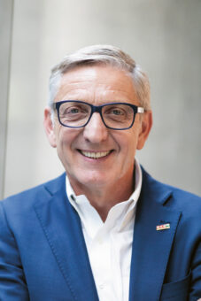 Ein lächelnder älterer Mann mit grauem Haar und Brille, der eine blaue Anzugsjacke und ein weißes Hemd trägt, steht vor einem unscharfen neutralen Hintergrund. Er trägt eine kleine Anstecknadel am Revers seines Jacketts.