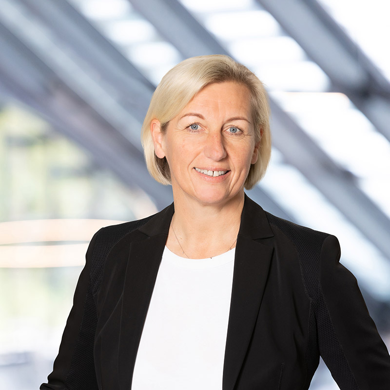 Eine Frau mit kurzen blonden Haaren, die einen schwarzen Blazer und ein weißes Top trägt, steht lächelnd vor einem unscharfen modernen architektonischen Hintergrund.