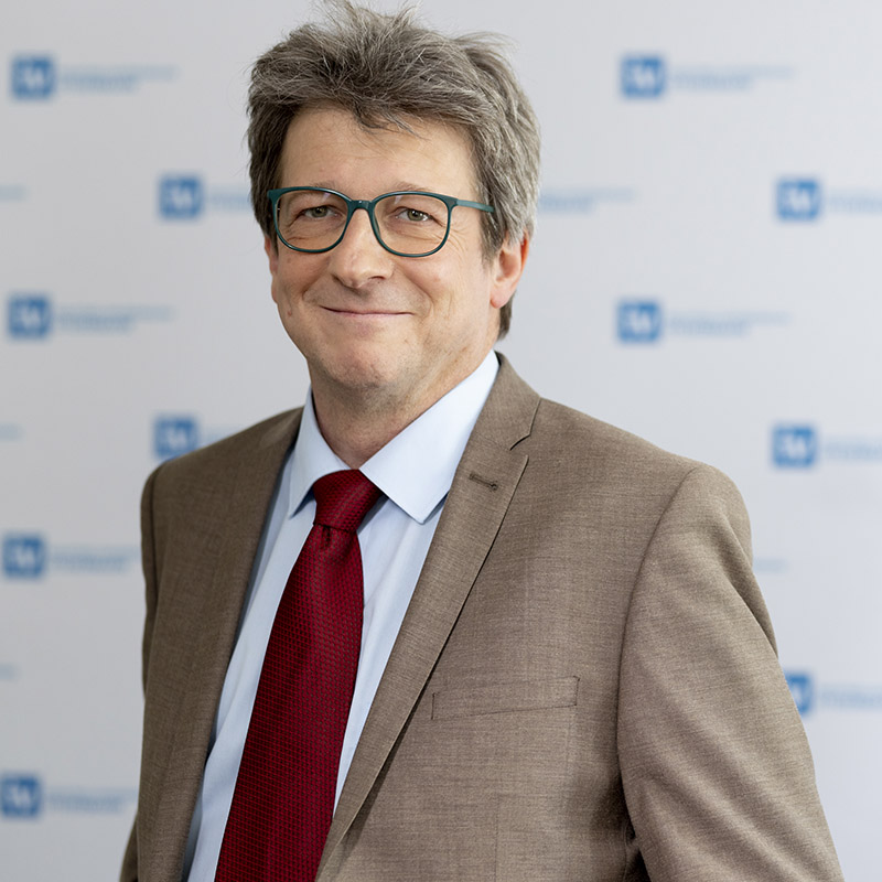 Ein Mann mit grauem Haar, Brille und einem braunen Anzug mit roter Krawatte lächelt in die Kamera. Im Hintergrund ist eine weiße Wand mit blauen und weißen Logos zu sehen.