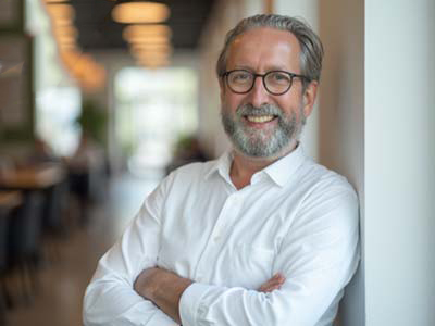 Ein lächelnder Mann mit Brille und Bart, der ein weißes Hemd trägt, steht mit verschränkten Armen an eine weiße Wand gelehnt in einem hellen, modernen Innenraum mit verschwommenen Lichtern im Hintergrund.